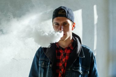 Vape genç. İlkbaharda sokakta elektronik sigara içen bir şapkalı genç yakışıklı adamın portresi. Sağlık için zararlı kötü alışkanlık. Vaping aktivitesi. Yakın çekim.