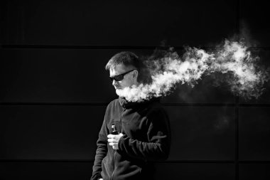 Vape adam. Güneş gözlüğü olan yetişkin bir sakallı adam, dışarıda güneşli bir günde elektronik sigara içiyor. Sağlık için zararlı kötü alışkanlık. Vaping aclivity. Siyah beyaz.