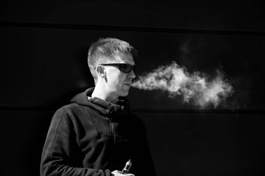 Vape adam. Güneş gözlüğü olan yetişkin bir sakallı adam, dışarıda güneşli bir günde elektronik sigara içiyor. Sağlık için zararlı kötü alışkanlık. Vaping aclivity. Siyah beyaz.