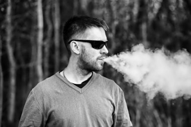 Vape adam. Gözlüklü bir yetişkin beyaz sakallı adam güneşli bir gün ormanda dışarıda bir elektronik sigara içiyor. Sağlık için zararlı kötü alışkanlık. Siyah beyaz.