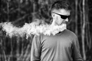 Vape adam. Gözlüklü bir yetişkin beyaz sakallı adam güneşli bir gün ormanda dışarıda bir elektronik sigara içiyor. Sağlık için zararlı kötü alışkanlık. Siyah beyaz.