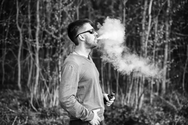 Vape man. An adult white bearded man in glasses smokes an electronic cigarette outside in the forest in sunny day. Bad habit that is harmful to health. Black and white.