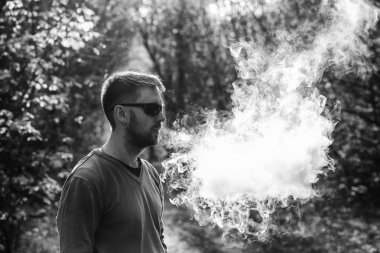 Vape adam. Güneş gözlüğü bir yetişkin beyaz sakallı adam güneşli bir gün ormanda dışarıda bir elektronik sigara içiyor. Sağlık için zararlı kötü alışkanlık. Siyah beyaz.