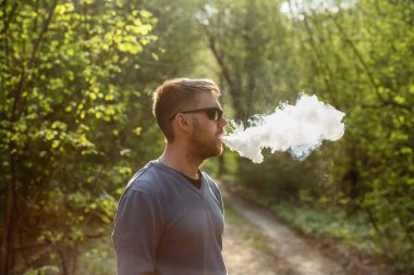 Vape adam. Güneş gözlüğü bir yetişkin beyaz sakallı adam güneşli bir gün ormanda dışarıda bir elektronik sigara içiyor. Sağlık için zararlı kötü alışkanlık.