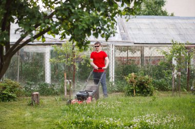 Kırmızı tişörtlü ve güneş gözlüklü genç adam güneşli bir yaz gününde köyün arka bahçesindeki çimenleri biçer..