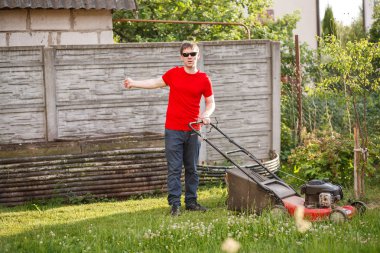 Kırmızı tişörtlü ve güneş gözlüklü genç adam güneşli bir yaz gününde köyün arka bahçesindeki çimenleri biçer..