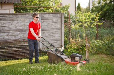 Kırmızı tişörtlü ve güneş gözlüklü genç adam güneşli bir yaz gününde köyün arka bahçesindeki çimenleri biçer..