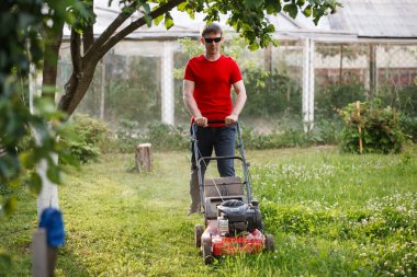 Kırmızı tişörtlü ve güneş gözlüklü genç adam güneşli bir yaz gününde köyün arka bahçesindeki çimenleri biçer..