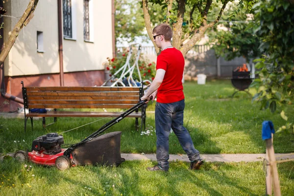 Kırmızı tişörtlü ve güneş gözlüklü genç adam güneşli bir yaz gününde köyün arka bahçesindeki çimenleri biçer..