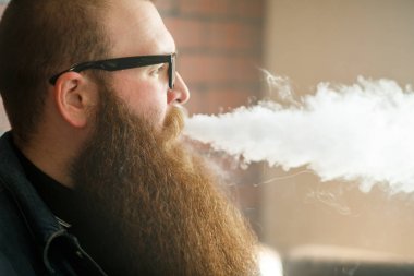 Vape sakallı adam. Gözlüklü çok uzun sakallı yetişkin bir adam barda elektronik sigara içiyor. Sağlık için zararlı kötü alışkanlık. Vaping etkinliği.