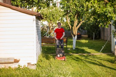 Kırmızı tişörtlü ve güneş gözlüklü genç adam güneşli bir yaz gününde köyün arka bahçesindeki çimenleri biçer. Bahçende çalış..