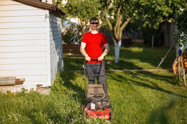 Kırmızı tişörtlü ve güneş gözlüklü genç adam güneşli bir yaz gününde köyün arka bahçesindeki çimenleri biçer. Bahçende çalış..