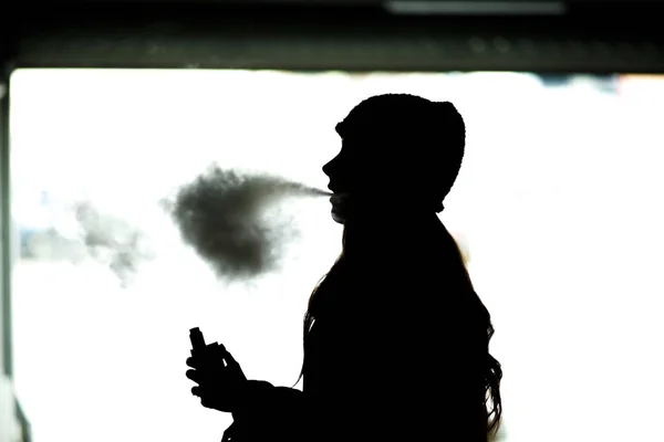 Vape genç. Elektronik sigara içen şapkalı genç bir kızın silueti. Sağlığa zararlı zararlı alışkanlık. Vaping etkinliği.