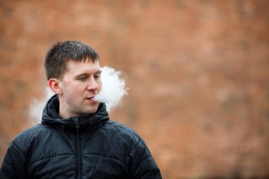 Vape adam. Siyah ceket sigara ve sonbahar içinde bulanık turuncu arka plan zıt bir elektronik sigara buhar dışarı izin genç Kafkas Beyaz adam. Kötü alışkanlık. Vaping aktivitesi. Yaşam tarzı.