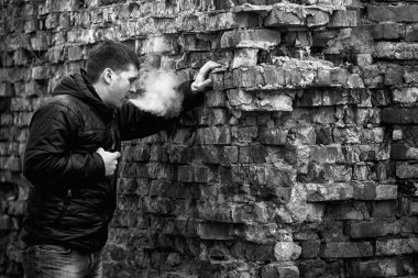 Vape adam. Siyah ceket sigara ve sonbahar eski yıkılan tuğla duvar yakın bir elektronik sigara buhar dışarı izin genç Kafkas Beyaz adam. Kötü alışkanlık. Vaping aktivitesi. Siyah beyaz