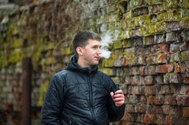 Vape adam. Siyah ceket sigara ve sonbahar eski yıkılan tuğla duvar yakın bir elektronik sigara buhar dışarı izin genç Kafkas Beyaz adam. Kötü alışkanlık. Vaping aktivitesi. Yaşam tarzı.