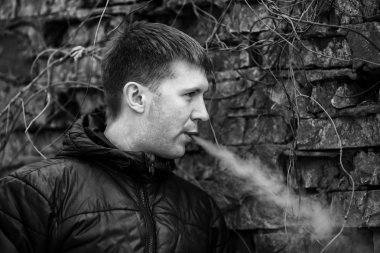 Vape adam. Siyah ceket sigara ve sonbahar eski yıkılan tuğla duvar yakın bir elektronik sigara buhar dışarı izin genç Kafkas Beyaz adam. Kötü alışkanlık. Vaping aktivitesi. Siyah beyaz