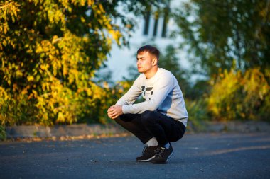 Sonbaharda gün batımında bir sokakta yeşil ağaçların önünde çömelmiş rahat giysiler içinde genç yakışıklı beyaz beyaz beyaz adam.