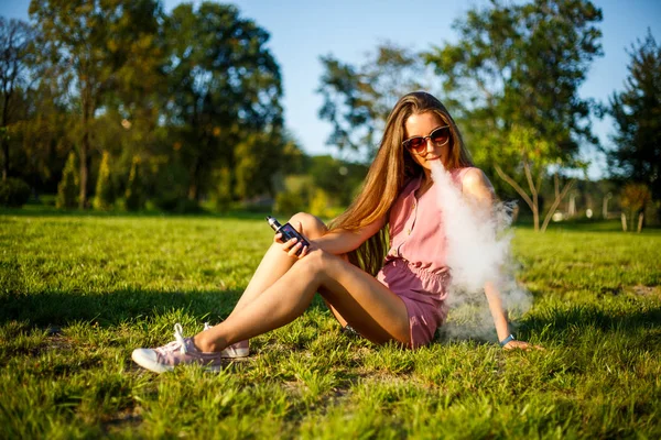 Vape genç. Pembe tulum ve güneş gözlüğü genç oldukça sportif kafkas kız yaz güneşli bir günde parkta elektronik sigara içiyor. Kötü alışkanlık. Vaping etkinliği.