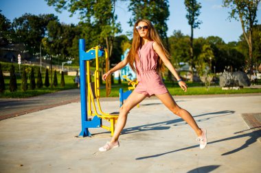 Pembe tulum ve güneş gözlüğü genç oldukça sportif kafkas kız yaz güneşli bir günde parkta sokak sporları alanında atlama. Sokak sporları zemininde fitness.