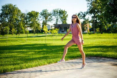 Genç. Pembe tulum ve güneş gözlüğü genç oldukça sportif kafkas kız yaz aylarında güneşli bir günde parkta atlama.