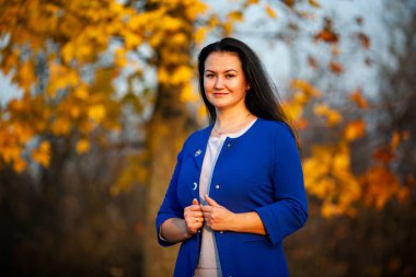 Altın sonbahar. Gün batımında şehir parkında mavi bir hırka güzel esmer beyaz kadın. Sarı yapraklar. Artı boyutlu model.