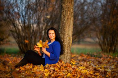 Altın sonbahar. Gün batımında şehir parkında mavi bir hırka güzel esmer beyaz kadın. Sarı yapraklar. Artı boyutlu model.