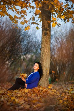 Altın sonbahar. Gün batımında şehir parkında mavi bir hırka güzel esmer beyaz kadın. Sarı yapraklar. Artı boyutlu model.