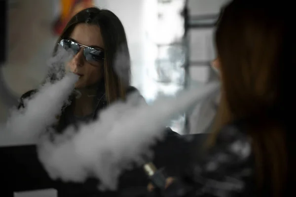 Sorunlu cildi olan Vape genç. Güneş gözlüğü genç sevimli kız portresi barda ayna karşısında elektronik sigara içen. Kötü alışkanlık. Vaping aktivitesi. Karanlık atış.
