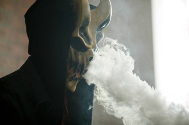 Şeytan maskeli vape adam ve kapalı bir elektronik sigara içen bir başlık. Buhar puffs. Sağlık için zararlı kötü alışkanlık. Vaping etkinliği.
