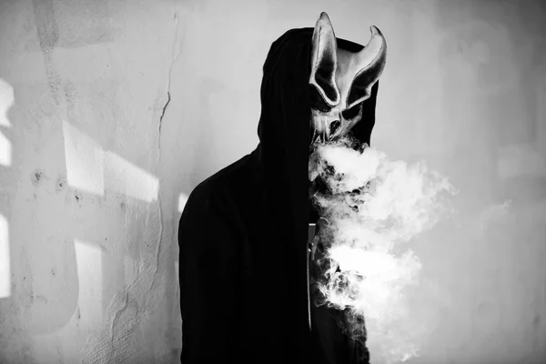Vape man in devil mask and a hood smoking an electronic cigarette outdoors. Puffs of steam. Bad habit that is harmful to health. Vaping activity. Black and white.