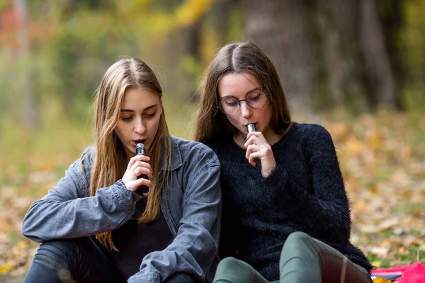 Vape lgbt gençleri. Biseksüel lezbiyen, beyaz, günlük kıyafetler giyen genç kızlar sonbahar akşamı parkta sokakta elektronik sigara yakarlar. Kötü alışkanlık..