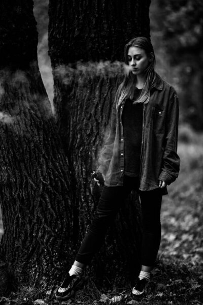 Vape teenager. Young white addict caucasian teenage girl in casual clothes vaping electronic cigarette with cannabis liquid near the tree in the forest in the autumn evening. Black and white.