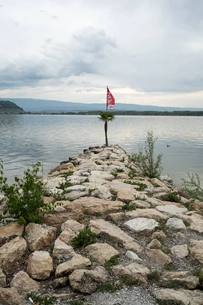 旗のポールと終わりとの背後にある美しい圧延丘と湖風景に旗で穏やかな湖へリードして岩の桟橋 — ストック写真