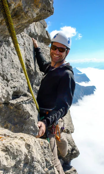 rock climber on a vertical rock face climbing and smiling above a sea of clouds in the valley below