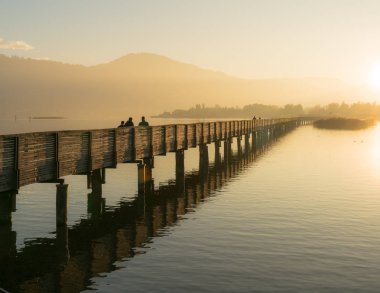 Uzun ahşap iskele ve altın akşam silhouette yayalar ve insanlar yürürken, insanlar ve arka planda dağlar ile hafif Rapperswil yakınındaki Zürih Gölü üzerinde tahta