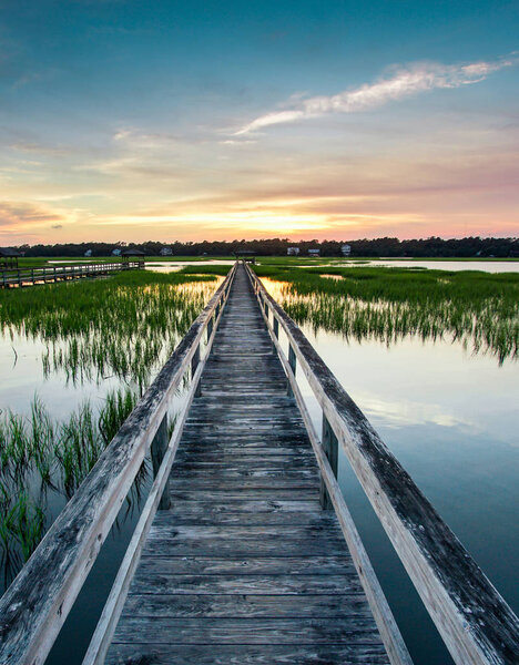 vertical view of a beautiful sunset over coastal waters with a very long wooden boardwalk pier in the center during a colorful summer sunset under an expressive sky with reflections in the water and marsh grass in the foreground
