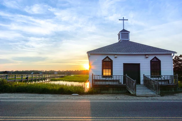 small white wooden chapel on the water on the coast during a colorful summer sunset with illuminated golden windows and a setting sun under a blue sky with soft clouds