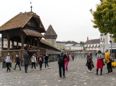 Lucerne, LU / Switzerland - November 9, 2018: tourists visit the famous Swiss city of Lucerne and take pictures of themselves with the Chapel Bridge  behind them as they stand on the river banks of the Reuss clipart