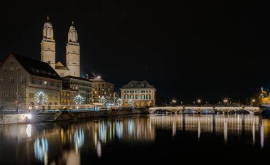 Zürih cityscape Limmat Nehri ile gece ve Grossmuenster Katedrali ve ışıklar ön planda yansımaları ile Köprü