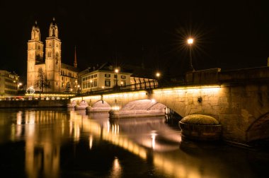 Zürih cityscape Limmat Nehri ile gece ve Grossmuenster Katedrali ve ışıklar ön planda yansımaları ile Köprü