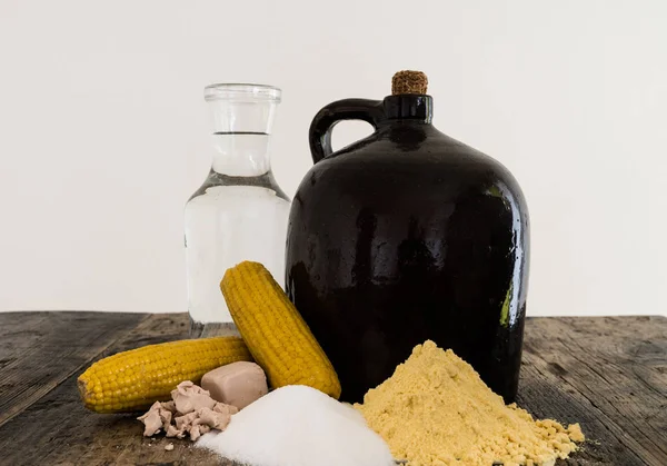 A clay half gallon jug with cob stopper and the ingredients for making moonshine corn liquor on a rustic wooden table
