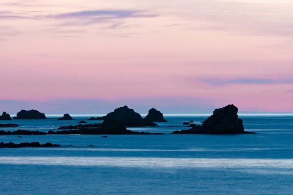 Puesta de sol en la playa con un océano tranquilo y rocas y arrecifes bajo un cielo lila púrpura — Foto de Stock