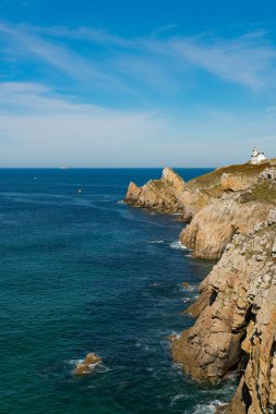 Derin mavi okyanusları ve beyaz bir deniz feneri olan sarp kayalık bir sahil şeridi.