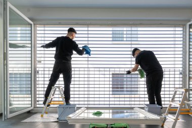 İki profesyonel erkek temizlikçinin yatay görüntüsü mikro fiber kumaşlarla dolu büyük bir pencerenin güneşliklerini temizliyor.