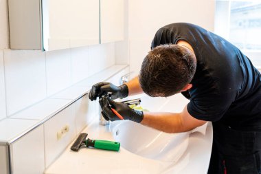 Kireç ve lekeleri temizlemek için banyo lavabosunu kazıyıcı bıçak ve mikro fiber bezle temizleyen bir adam.