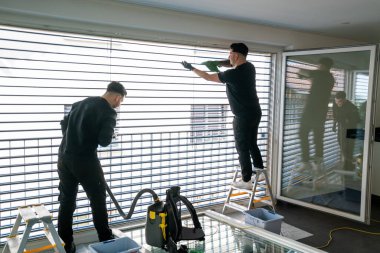 İki profesyonel erkek temizlikçinin yatay görüntüsü mikro fiber kumaşlarla dolu büyük bir pencerenin güneşliklerini temizliyor.