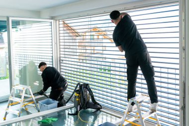 İki profesyonel erkek temizlikçinin yatay görüntüsü mikro fiber kumaşlarla dolu büyük bir pencerenin güneşliklerini temizliyor.