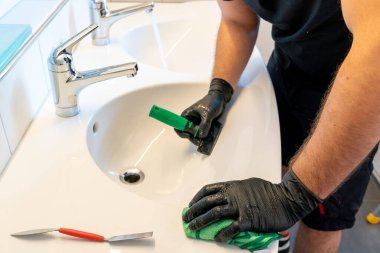 Profesyonel bir temizlikçi seramik banyo lavabosundan elmas bıçaklı kazıyıcı ve kumaşla toprak lekelerini temizliyor.