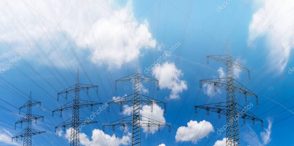 Several large metal high voltage lattice crosses transporting electricity under a blue sky with white clouds and light fog
